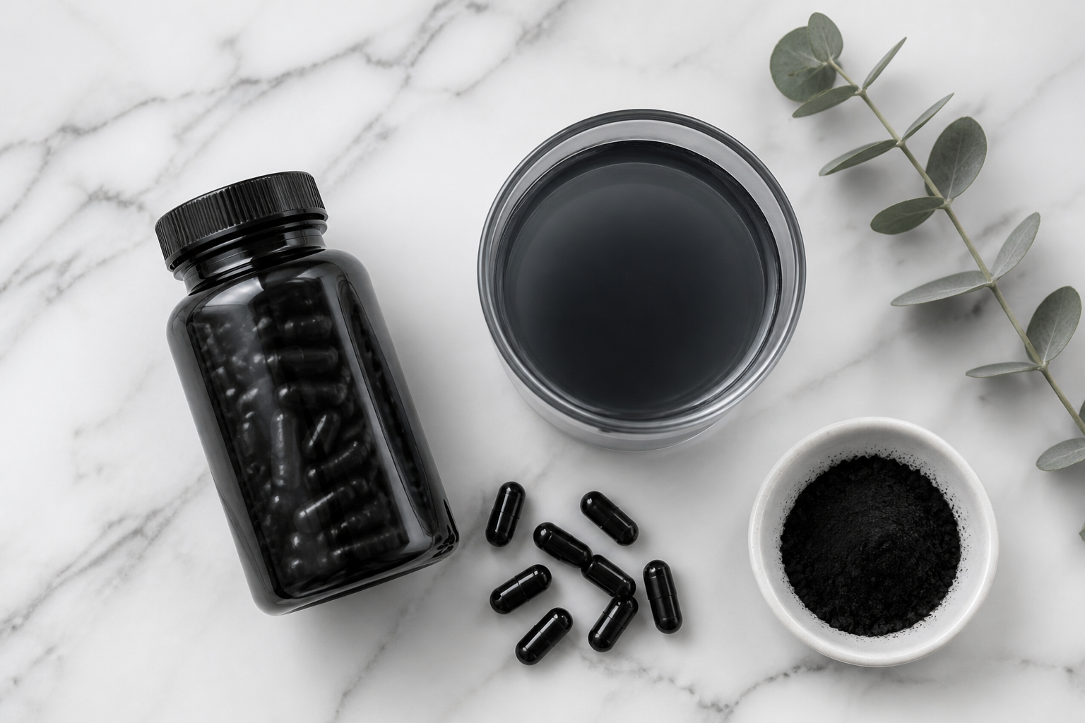 Flat lay of an activated charcoal capsule bottle beside a glass of charcoal water on a white marble surface